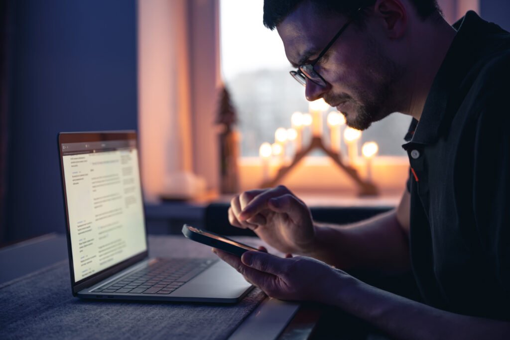 A man with a smartphone sits in front of a laptop late at night